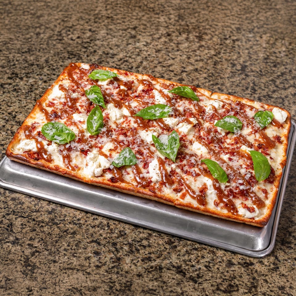 Rectangular cheesy pizza topped with crumbled meat, fresh basil, grated cheese, and sauce on a metal baking tray.